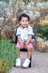 Toddler smiling at camera while sitting in mom’s lap with wildflowers and pond in background