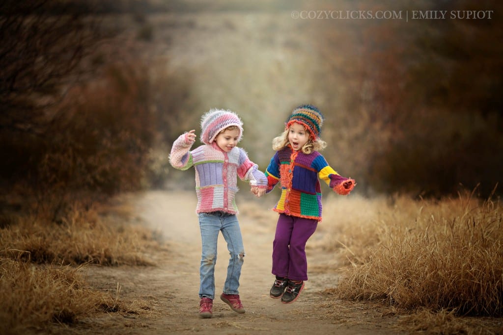 Two sisters jumping together in this picture wearing handmade sweaters and hats.